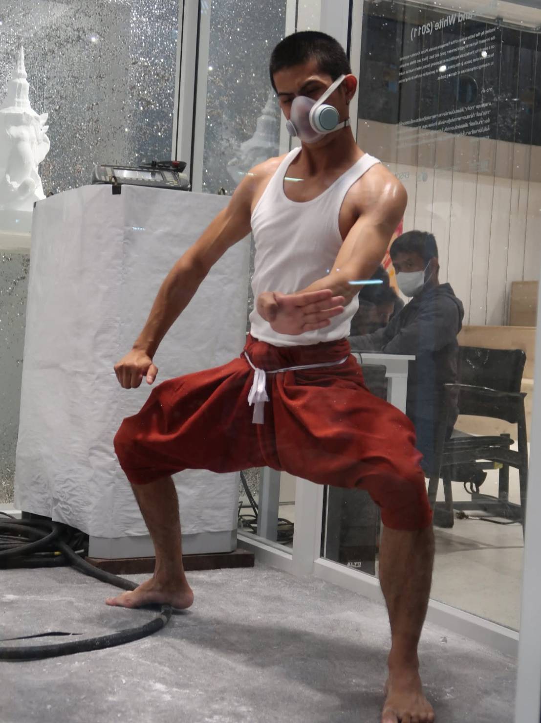 A male performance artist in a dynamic pose wearing a respirator and traditional red pants.