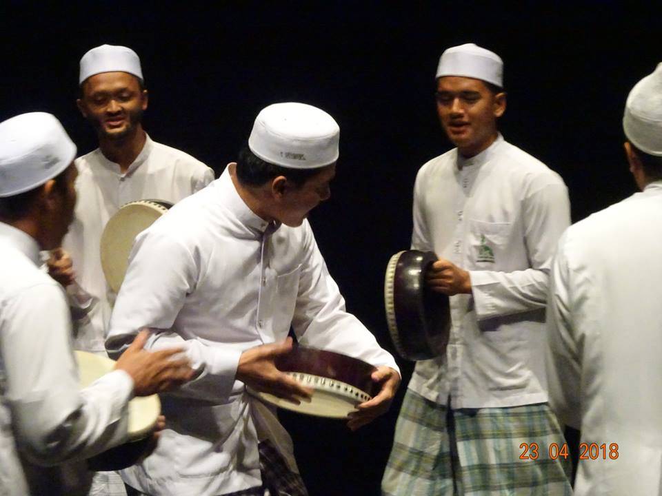 A group of men in traditional white clothing and hats playing hand drums during a performance.